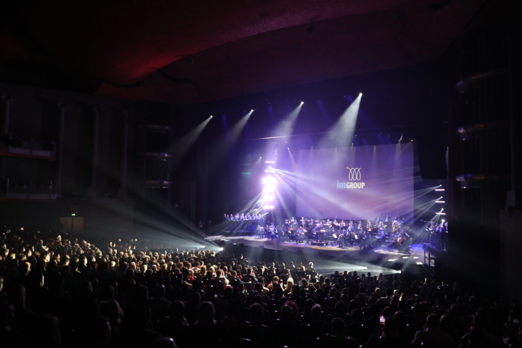 A large audience watches an orchestra perform on a stage with dramatic lighting. The background features a screen displaying the imGROUP logo.