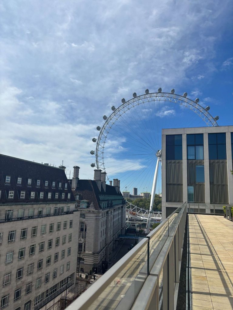 A large Ferris wheel, the London Eye, stands behind city buildings under a partly cloudy blue sky. A glass-railed walkway is on the right side of the image.