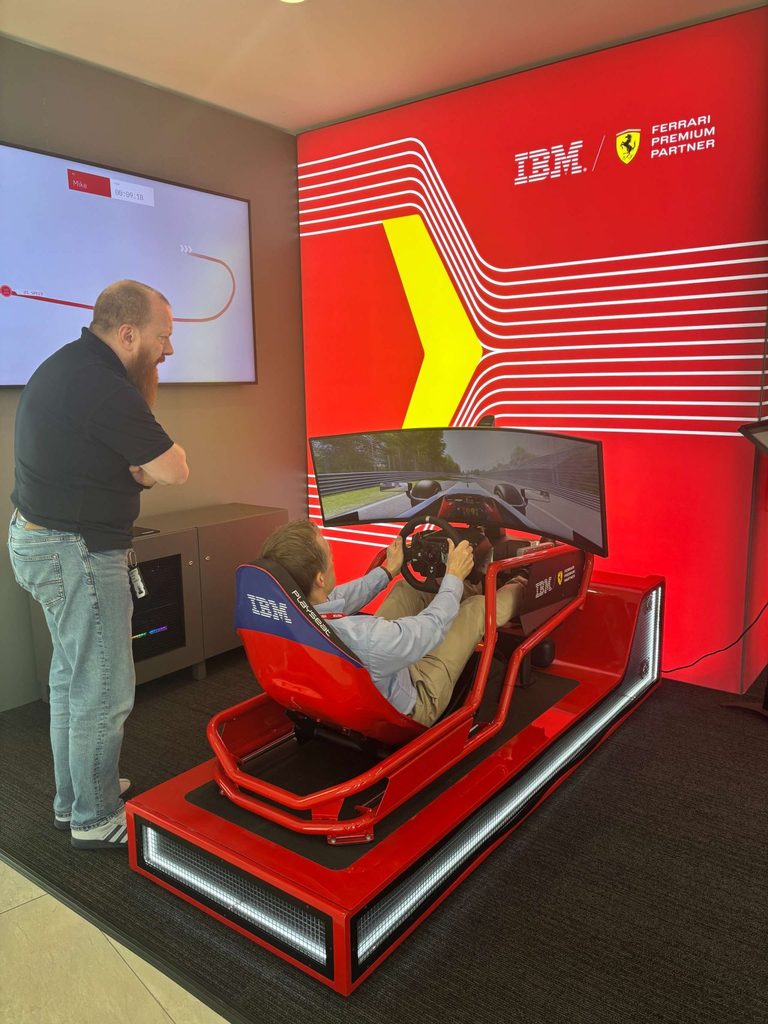 A man sits in an IBM-branded racing simulator, driving with pedals and a steering wheel, whilst another man stands beside him. A large red display behind them shows IBM and Ferrari logos.