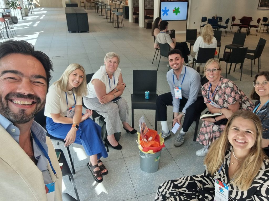 Seven smiling people sit in a circle indoors at a conference, some holding notebooks. Bright decorations are in a bucket in the centre. A presentation screen and more attendees are visible in the background.