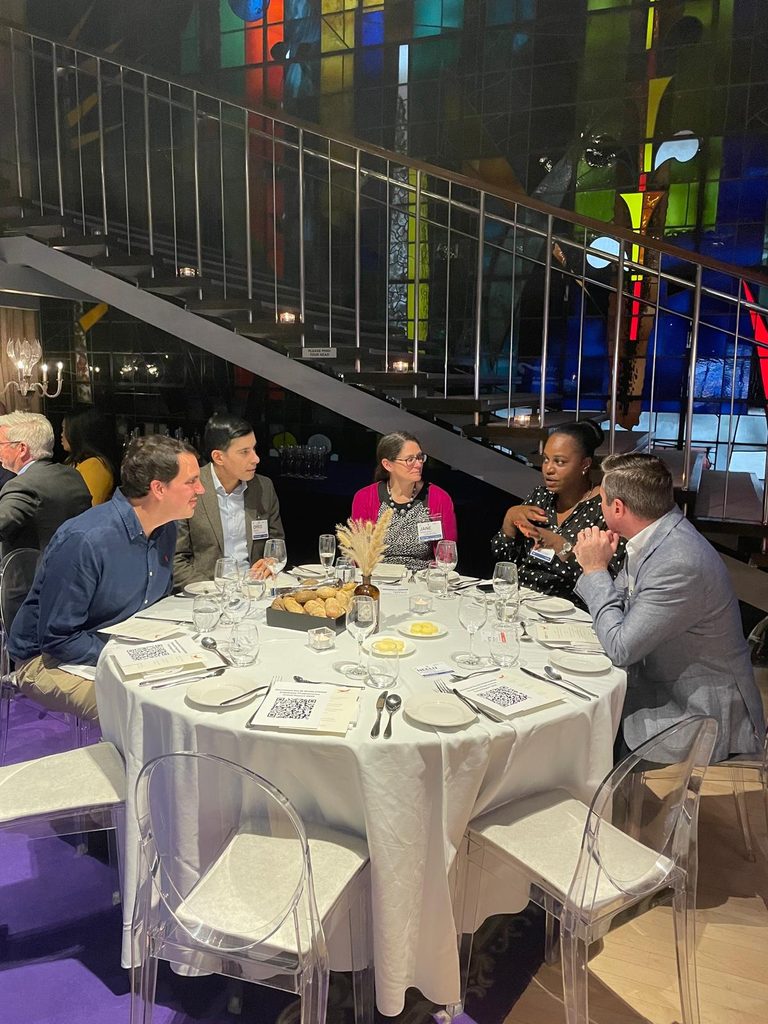 Five people sit around a round table set for dinner, engaged in conversation. The background features a staircase and colourful stained glass panels. The table has plates, glasses, bread, and a small floral centrepiece.
