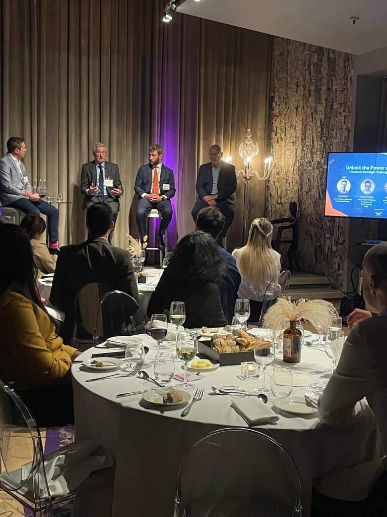 Four men in suits sit on a stage panel, speaking to an audience at a formal dinner event. A screen displays a presentation titled Unlock the Power. Attendees listen whilst seated at tables with food and wine.