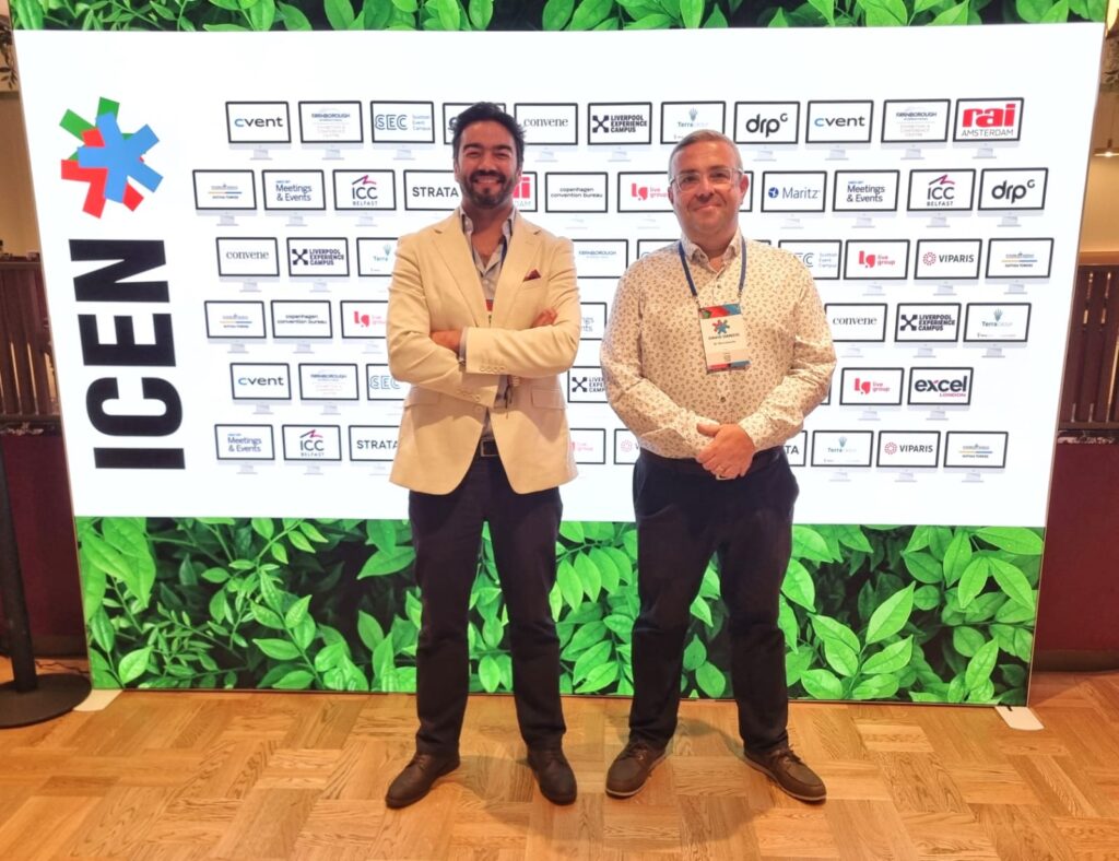 Two men stand smiling in front of a large display showing the ICEN logo and many sponsor logos on a green leafy background. Both wear conference badges and business casual attire.