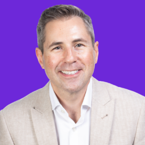 A smiling man wearing a white collared shirt and a beige blazer against a solid purple background.