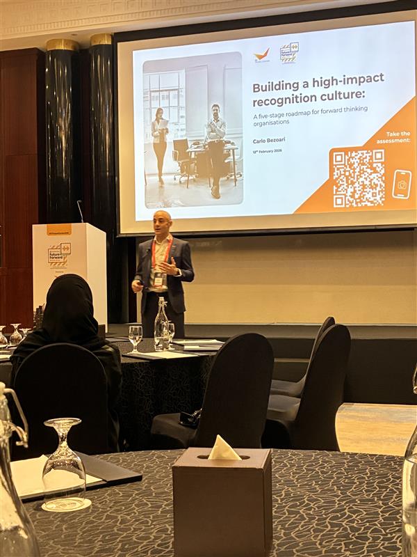 A speaker stands in front of a screen presenting Building a high-impact recognition culture to an audience at a conference. The audience is seated at tables with black chairs and glassware.