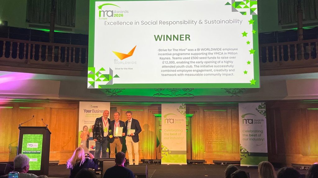 Three people stand on stage holding awards. A large screen above reads Excellence in Social Responsibility & Sustainability, WINNER, BI WORLDWIDE with event details and green décor in the background.