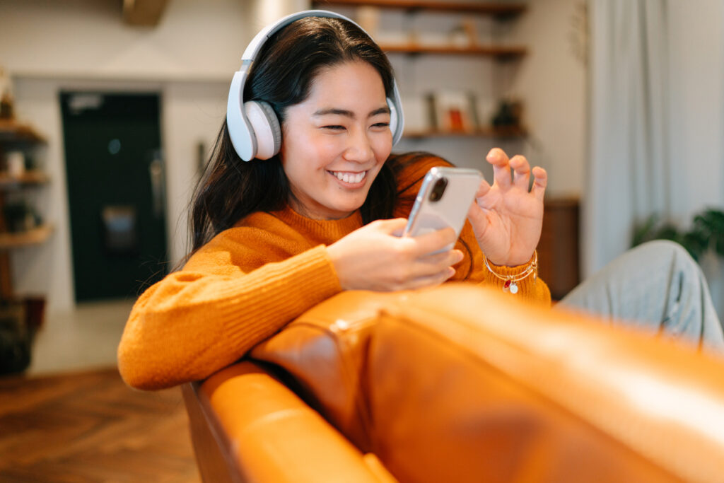 Một người phụ nữ đeo tai nghe và mặc áo len màu cam ngồi trên chiếc ghế dài màu nâu, mỉm cười và vẫy tay vào điện thoại thông minh trong cuộc gọi video trong phòng khách ấm cúng.