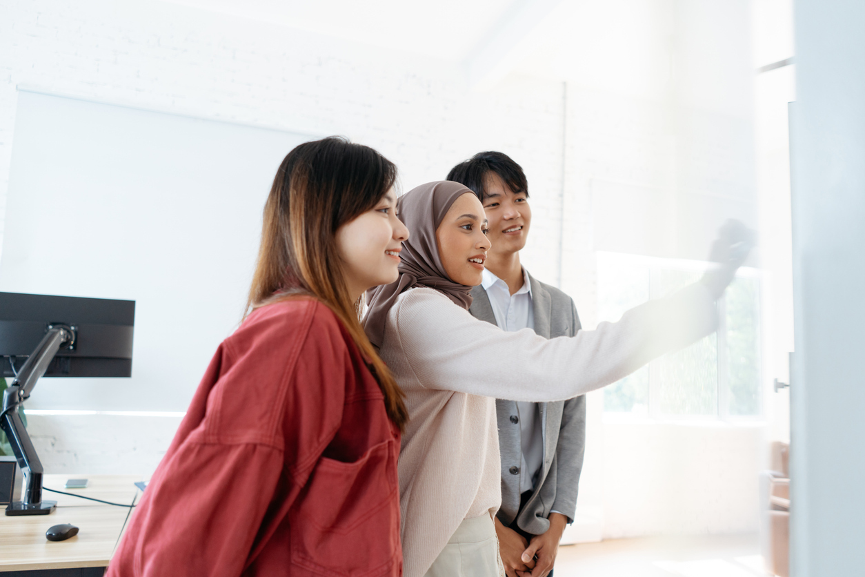 Ba chuyên gia trẻ, hai phụ nữ và một đàn ông, đứng cạnh nhau mỉm cười khi một người viết lên bảng trắng trong một văn phòng hiện đại, sáng sủa.