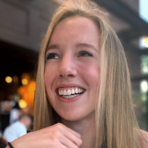 A woman with long blonde hair is smiling widely, looking upwards. The background is blurred, but appears to be an indoor setting with warm lighting.