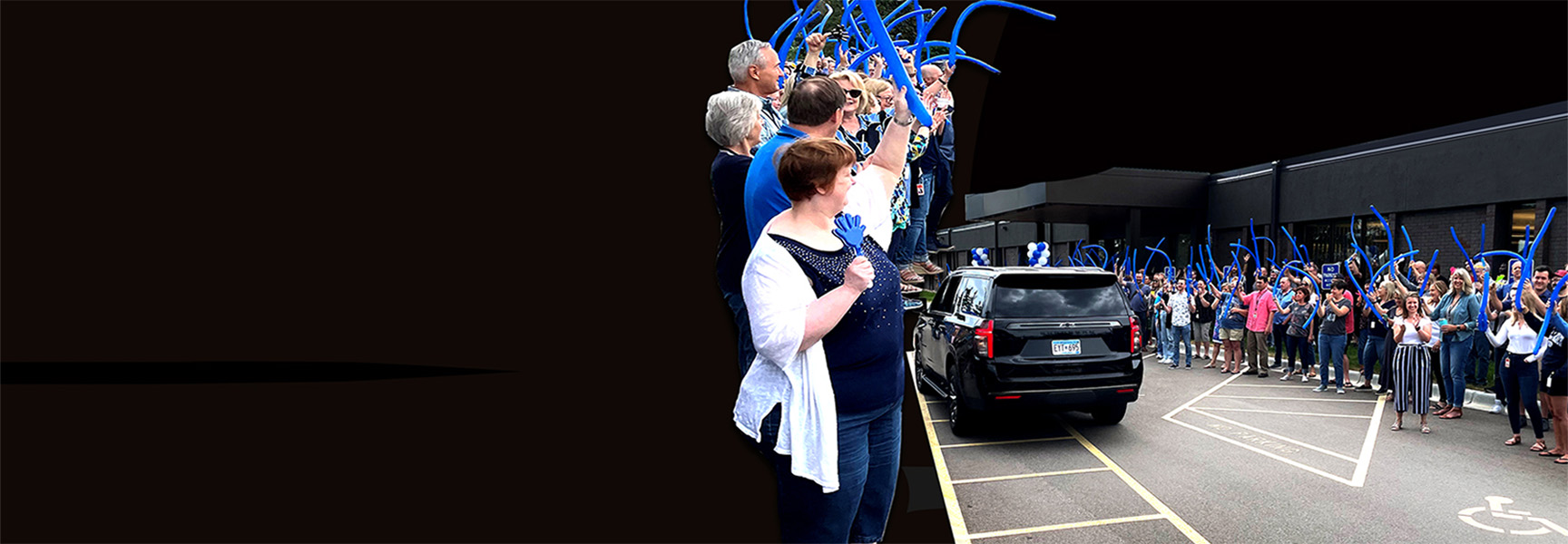 A crowd of people stands on either side of a road, some holding blue balloon-like objects, as a black car drives through. They appear to be cheering or celebrating as the car passes by.