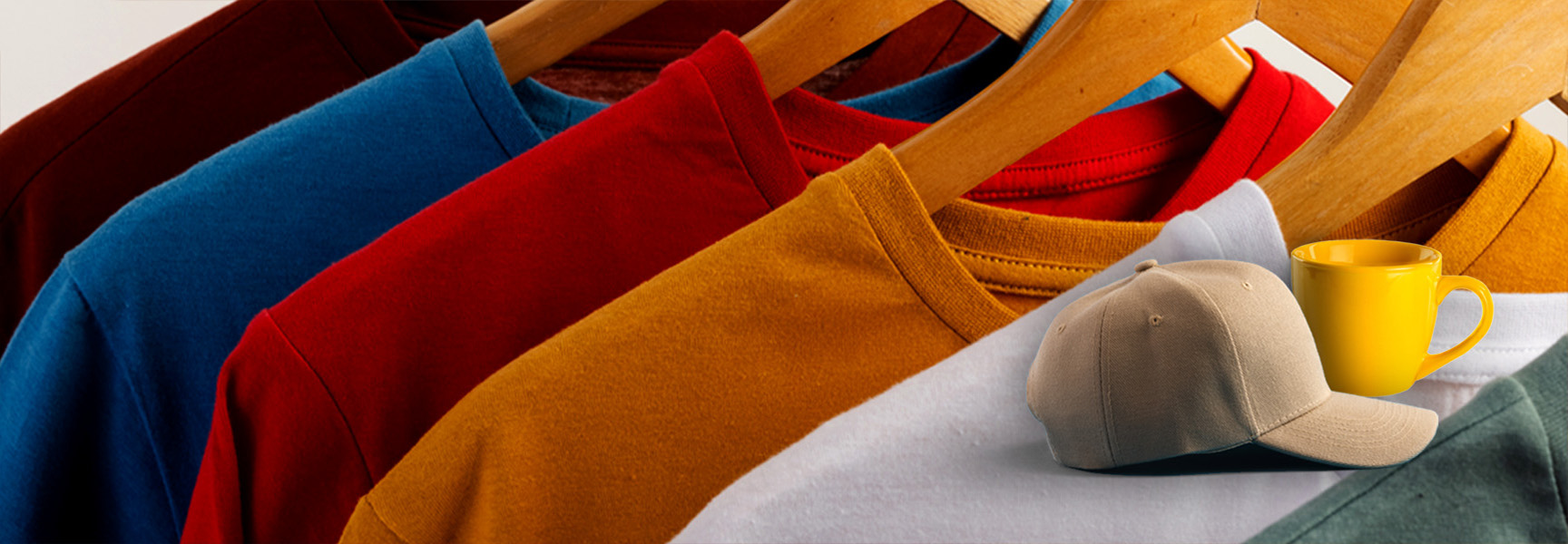 A row of colorful t-shirts hangs on wooden hangers. In front, a beige baseball cap and a yellow mug are placed against the shirts. The t-shirts include red, blue, orange, and white colors.