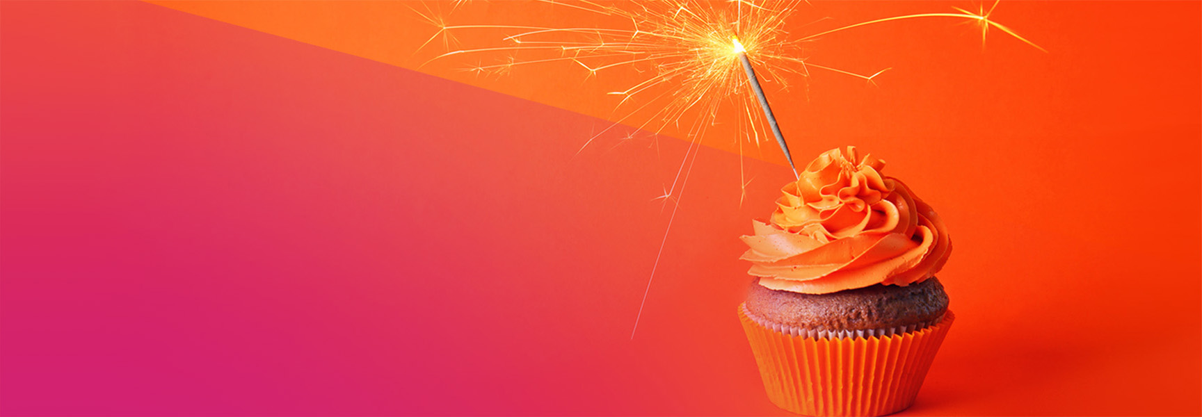 A cupcake with orange frosting sits in an orange wrapper. A lit sparkler is inserted into the frosting. The background is a gradient of orange and pink.