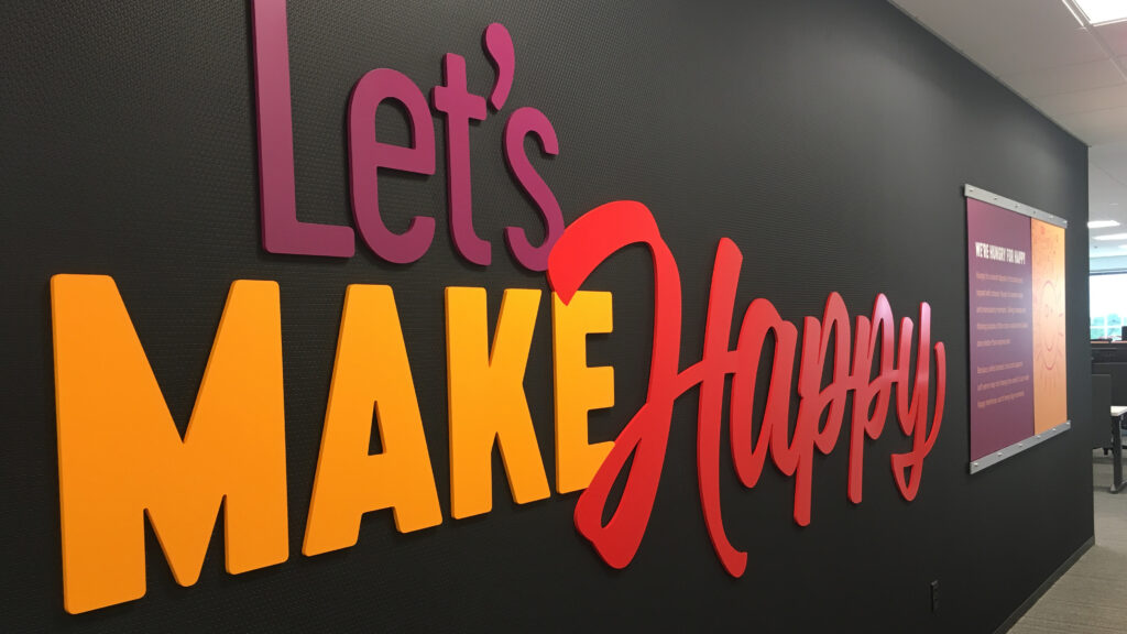 3D wall art in vibrant colors reads Lets MAKE Happy in an office hallway, with a purple, yellow, and red color scheme. Another sign with unreadable text hangs further down the hallway.