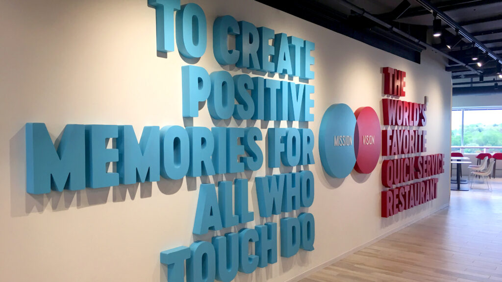 Large teal letters on a white wall read, “TO CREATE POSITIVE MEMORIES FOR ALL WHO TOUCH DQ,” alongside red letters stating, “THE WORLD’S FAVORITE QUICK-SERVICE RESTAURANT,” with “MISSION | VISION” in circles between them.