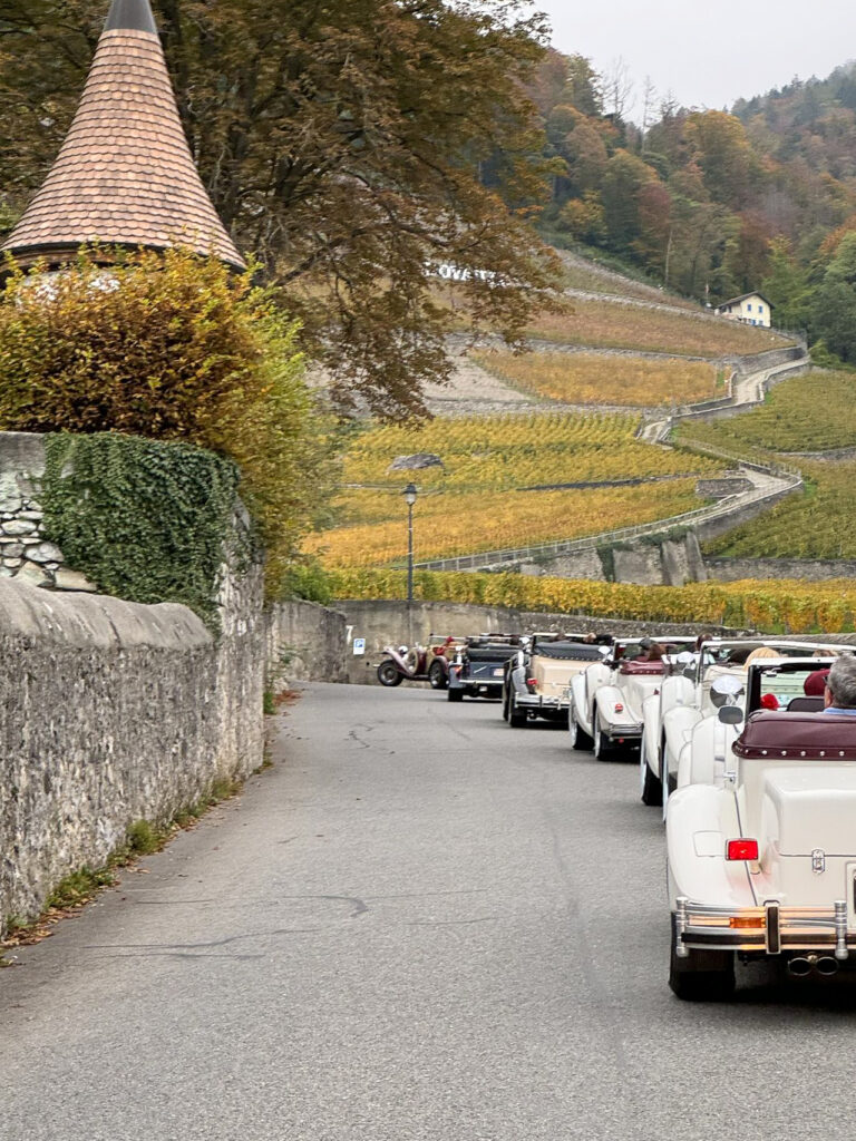 A line of vintage cars embarks on a group travel adventure along a narrow road bordered by stone walls. The road gracefully curves through a vineyard-covered hillside, with a medieval-style tower on the left and autumn foliage adorning the trees and vines.