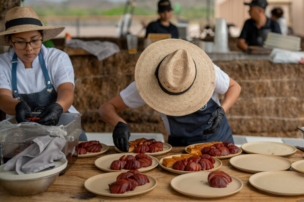 Two people wearing wide-brimmed hats and gloves are arranging sliced red fruit on beige plates at an outdoor food preparation area, a lively group travel scene. A stack of hay bales stands in the background with more travelers working enthusiastically.