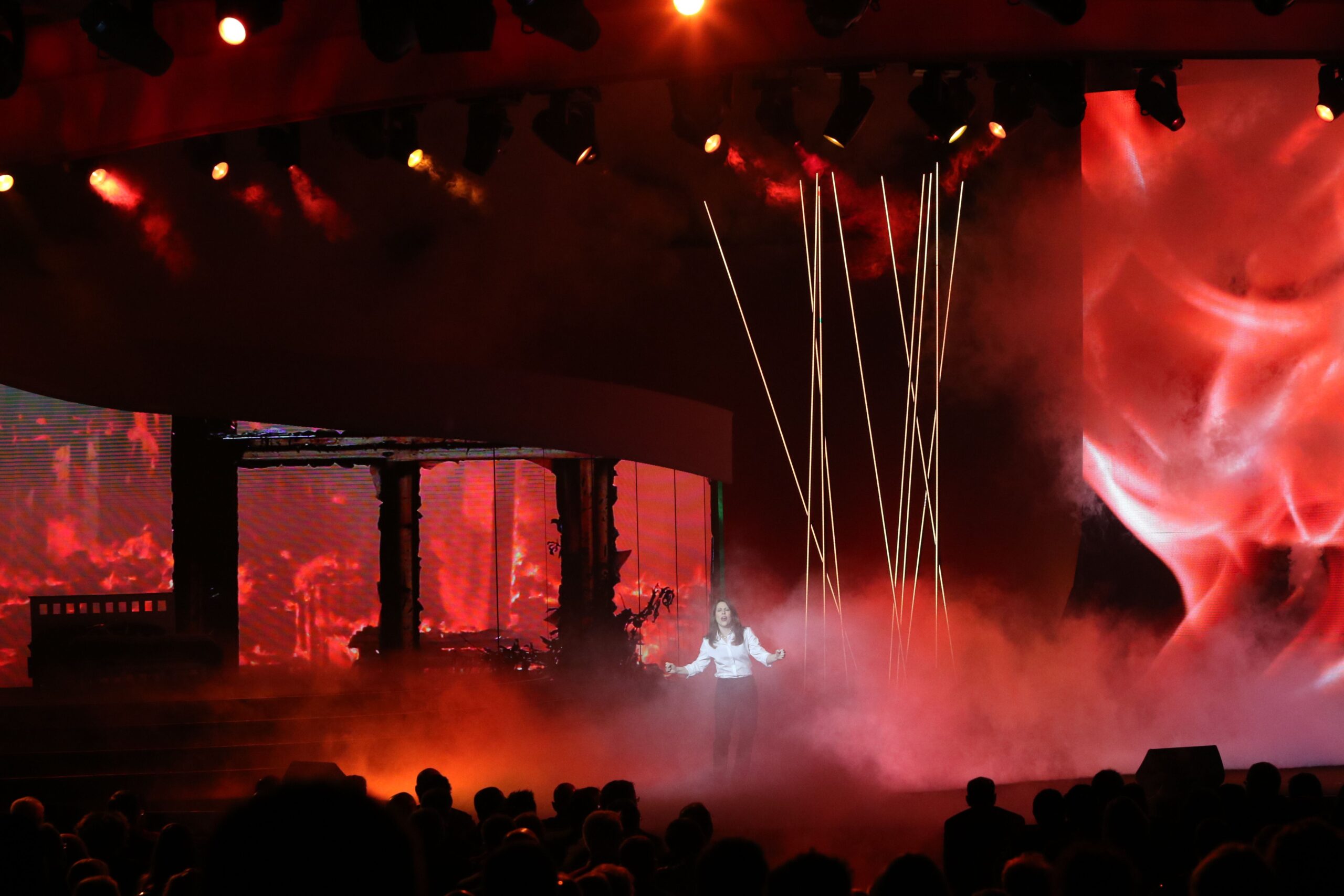 A performer on stage with dramatic red lighting and smoke effects, surrounded by vertical light beams. The audience is silhouetted in the foreground, and a large screen displays abstract visuals.