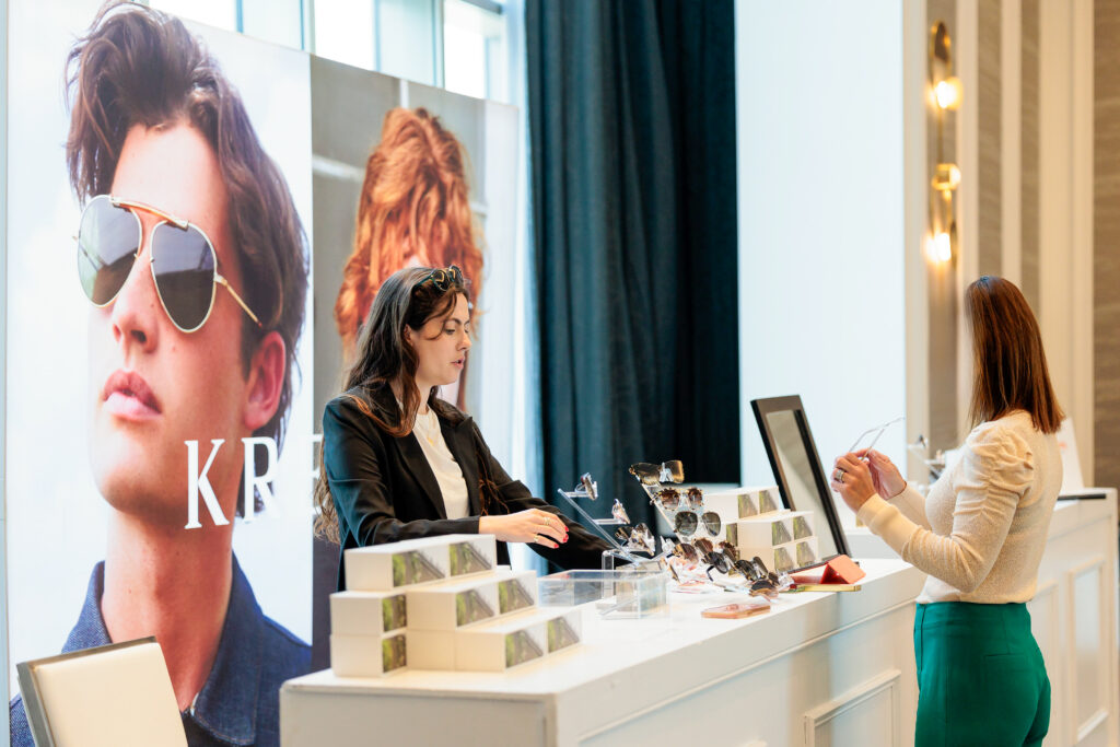 A woman, possibly planning a group travel adventure, stands at a sunglasses display counter conversing with a sales assistant. The counter features trendy sunglasses and promotional posters with models donning stylish shades in this bright, upscale store or showroom.