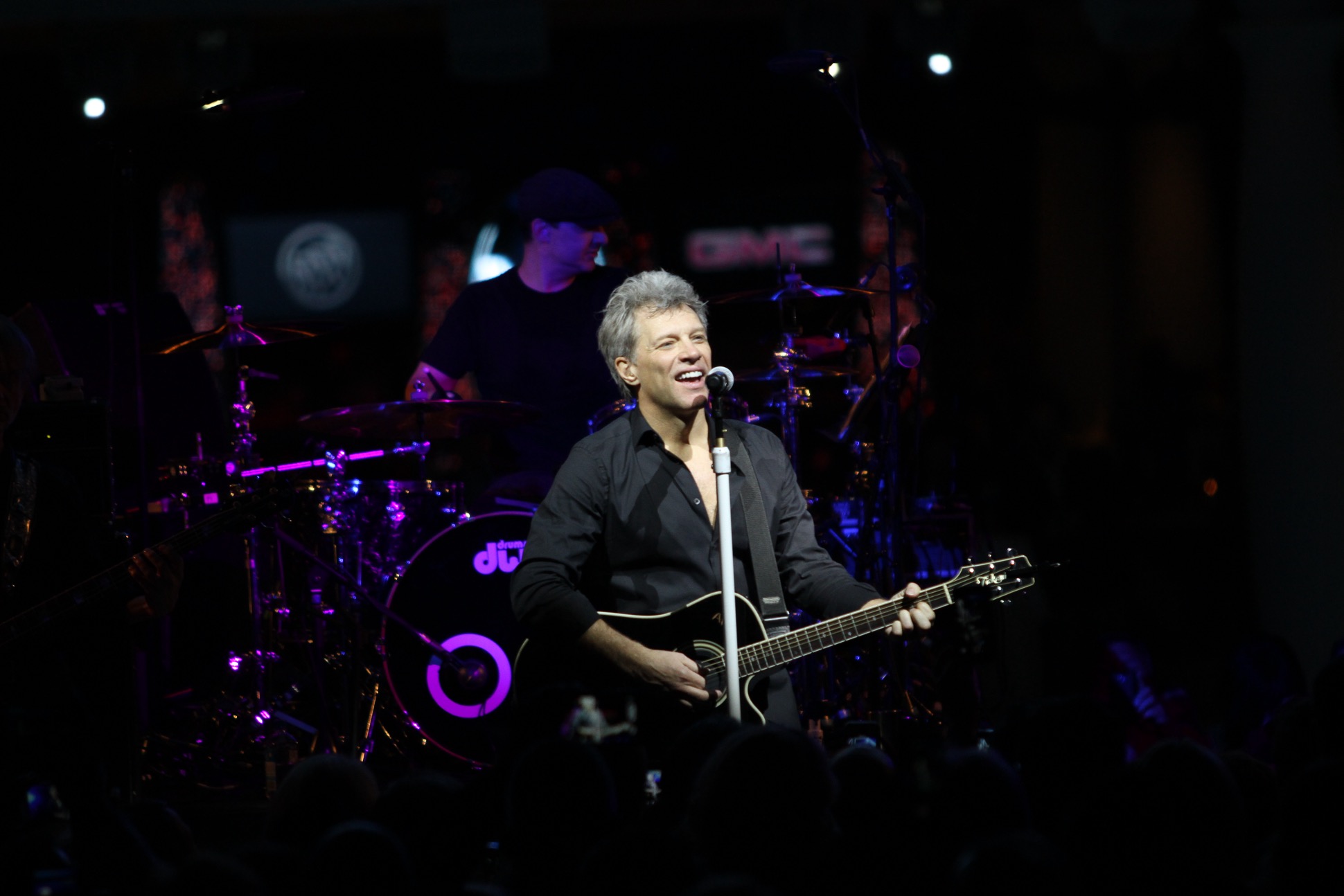 A musician with short gray hair performs on stage, playing a black acoustic guitar and singing into a microphone. A drummer is visible in the background, and the stage is illuminated with purple lighting.