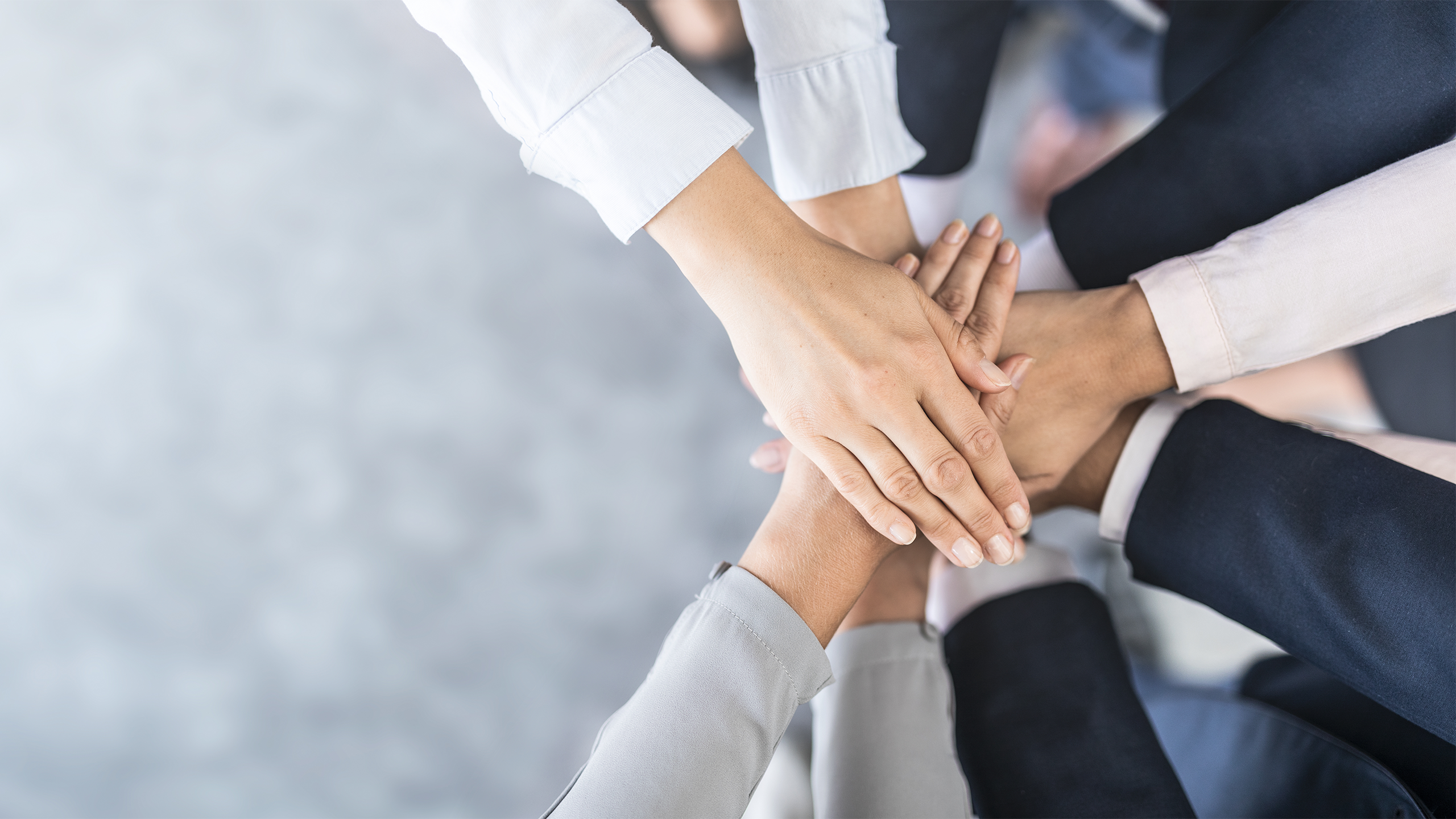 A group of people standing in a circle with their hands stacked on top of each other, symbolizing teamwork and unity. The background is blurred, highlighting the focus on the hands.
