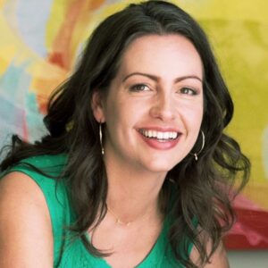 A woman with wavy dark hair, wearing a green sleeveless top and gold hoop earrings, smiles in front of a colorful abstract background.