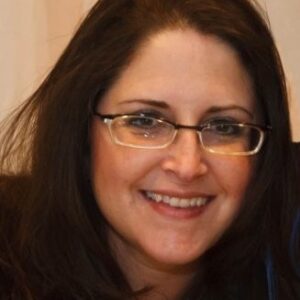 A woman with long brown hair and glasses smiles at the camera. She is wearing a dark top and is pictured against a softly lit, neutral background.