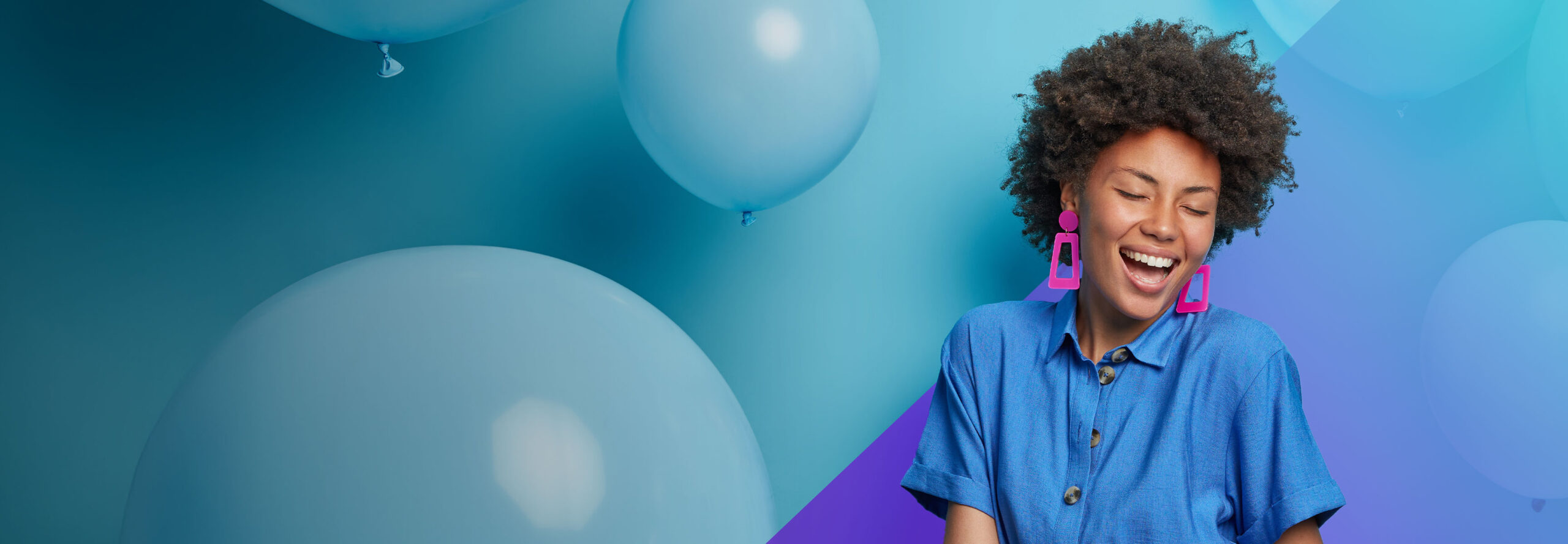 A joyful woman with curly hair, wearing a blue shirt and large pink earrings, smiles with her eyes closed in front of a blue background with large blue balloons.