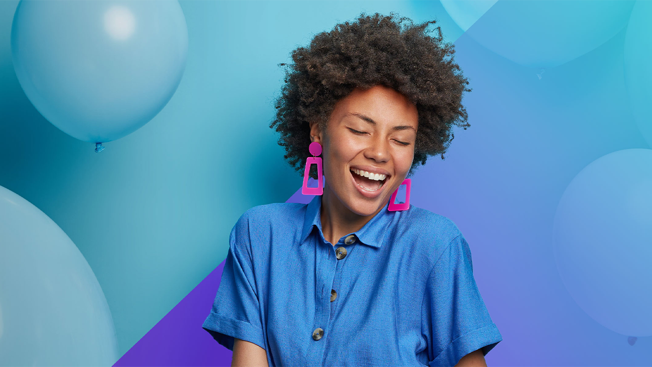 A woman with curly hair and bright pink earrings smiles joyfully with her eyes closed, wearing a blue shirt, standing in front of a blue and purple background with large balloons.