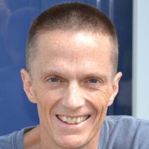 A middle-aged man with short brown hair and blue eyes smiles at the camera, wearing a gray shirt and standing in front of a blue background.