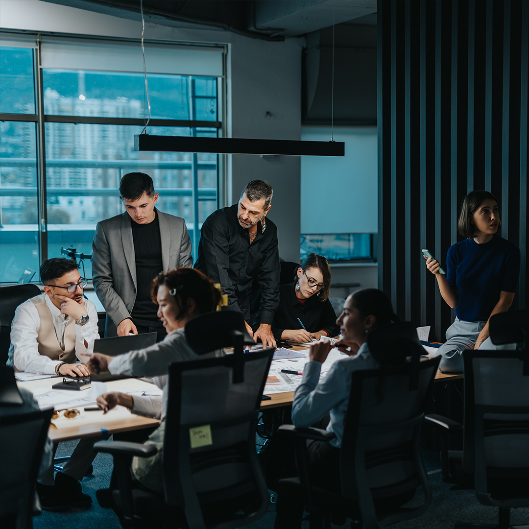 A group of professionals collaborate around a conference table in a modern office, discussing documents and ideas, with city buildings visible through large windows in the background.
