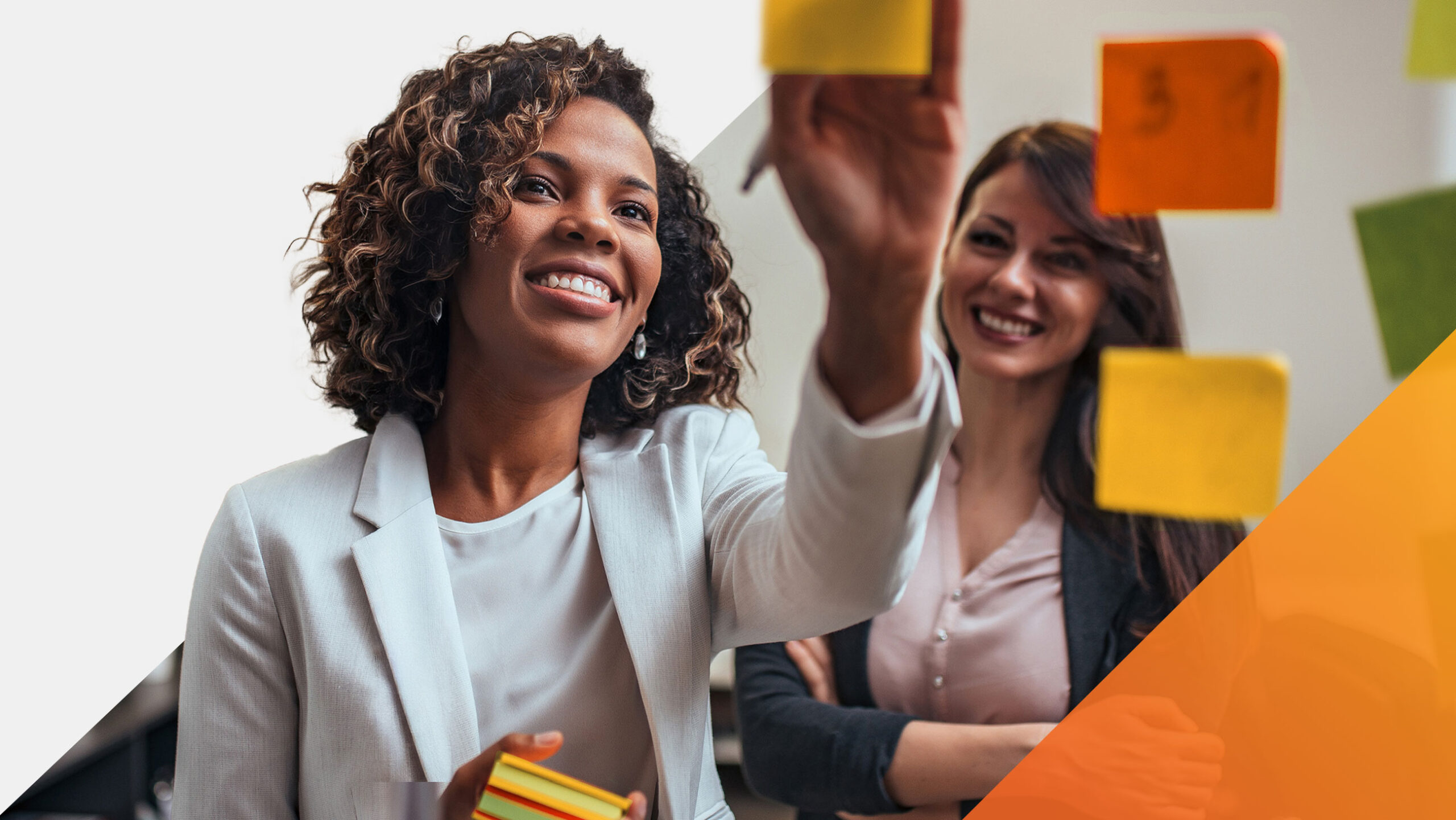 Two women smiling and placing colorful sticky notes on a glass wall, appearing engaged in a collaborative brainstorming or planning session in a modern office setting.
