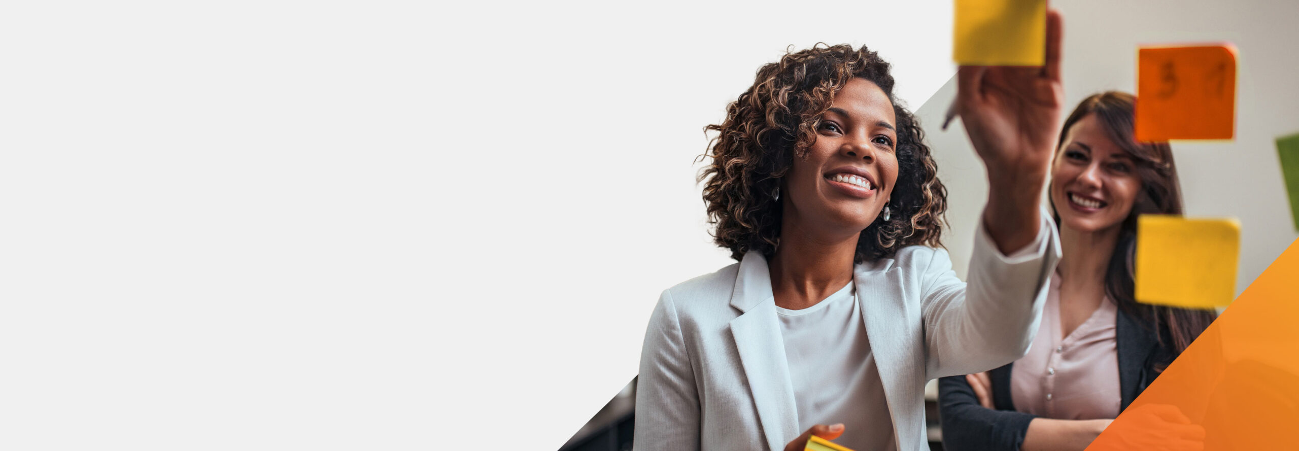 Two women, both smiling, stand together as one places colorful sticky notes on a glass board during a collaborative meeting. The background is mostly white, giving a bright and open feel.
