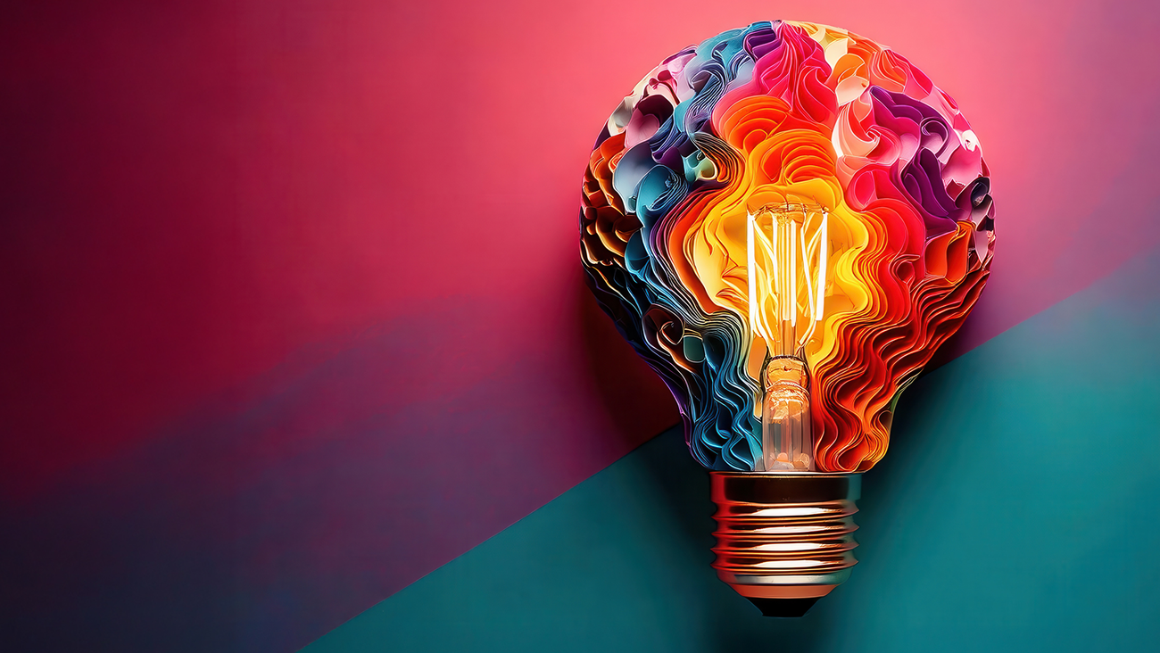 A colorful, artistic light bulb with intricate, layered paper-like patterns inside, glowing warmly against a vibrant red and teal background.