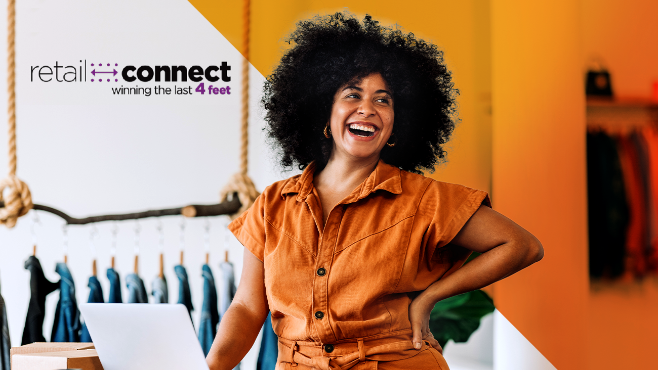 Smiling woman with curly hair stands by a laptop in a brightly lit store. The retail connect: winning the last 4 feet logo appears in the top left corner. Clothes hang in the blurred background.