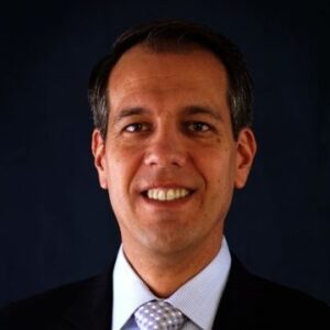 A man with short dark hair, wearing a dark suit, light blue shirt, and a light-patterned tie, smiles at the camera against a dark background.
