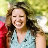 A woman with wavy, light brown hair is smiling broadly outdoors, wearing a sleeveless light blue top. The background is blurred greenery, suggesting a park or garden setting.