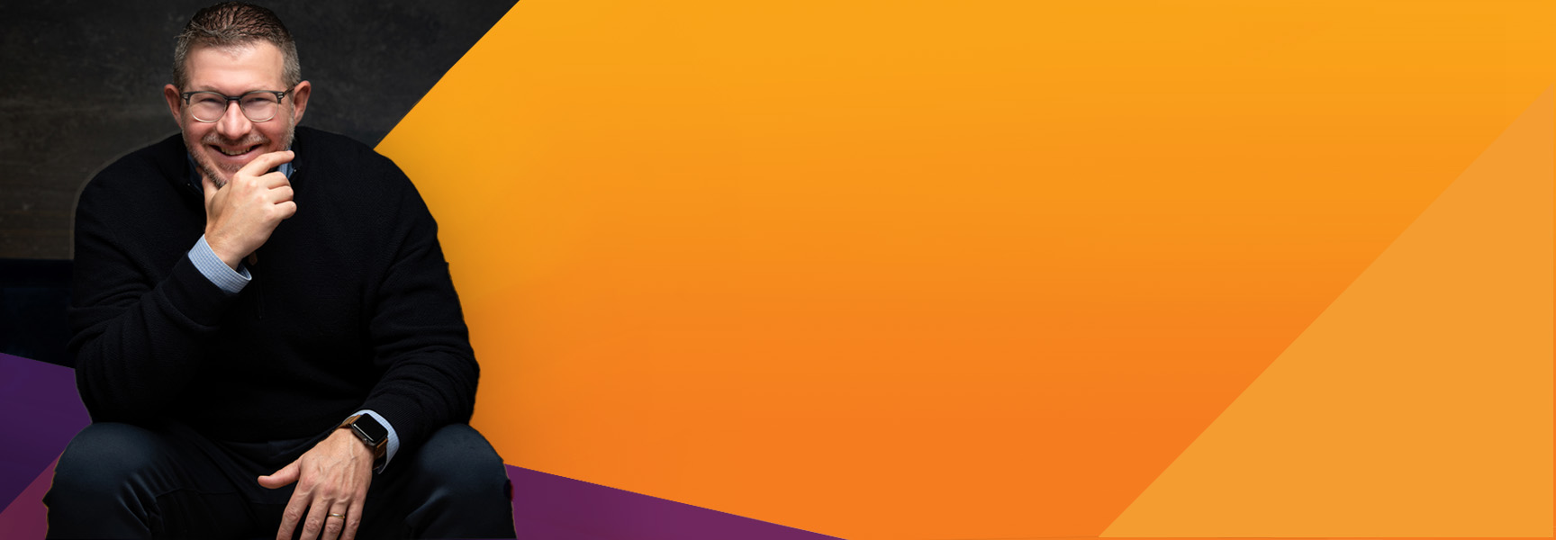 A smiling man with glasses and a beard, wearing a black sweater, sits with his hand on his chin against a background of yellow and orange geometric shapes.