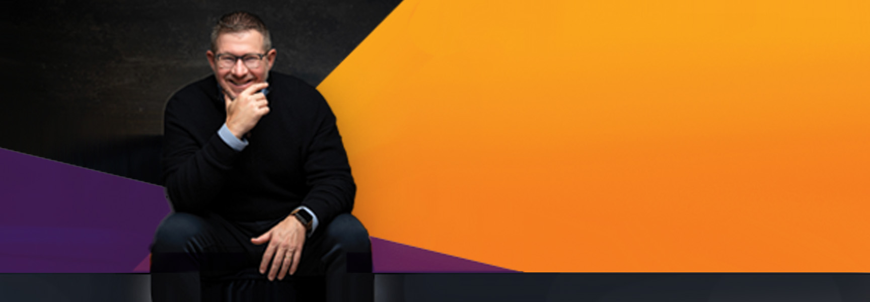 A smiling man wearing glasses and a black sweater sits against a background with geometric shapes in black, purple, and orange.