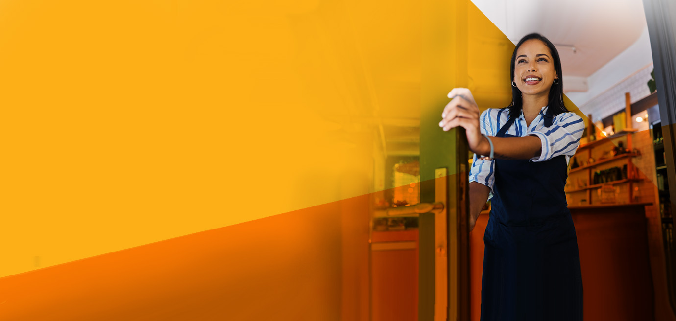 A smiling woman wearing an apron opens the door to a shop or cafe, standing in a warmly lit interior with shelves in the background. The left side of the image is overlaid with a large yellow geometric shape.
