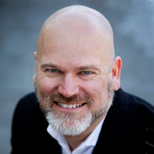 A smiling bald man with a beard and mustache, wearing a dark jacket and white collared shirt, posed against a blurred gray background.
