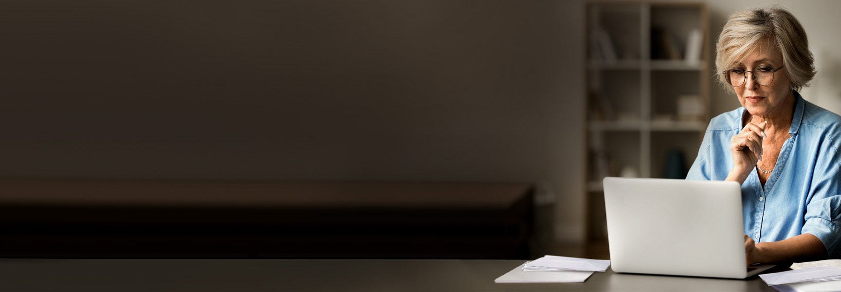 An older woman with glasses and short gray hair sits at a desk, looking thoughtfully at a laptop. Papers are on the desk, and a bookshelf is in the background. The left side of the image is mostly empty.