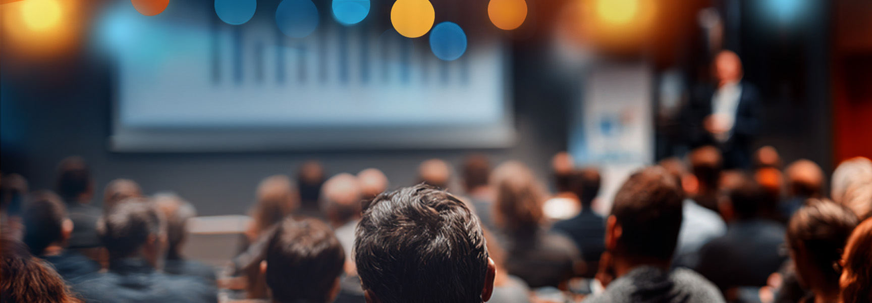 A blurred crowd sits facing a speaker and a large presentation screen with charts, under colorful, out-of-focus lights, suggesting a conference or seminar setting.