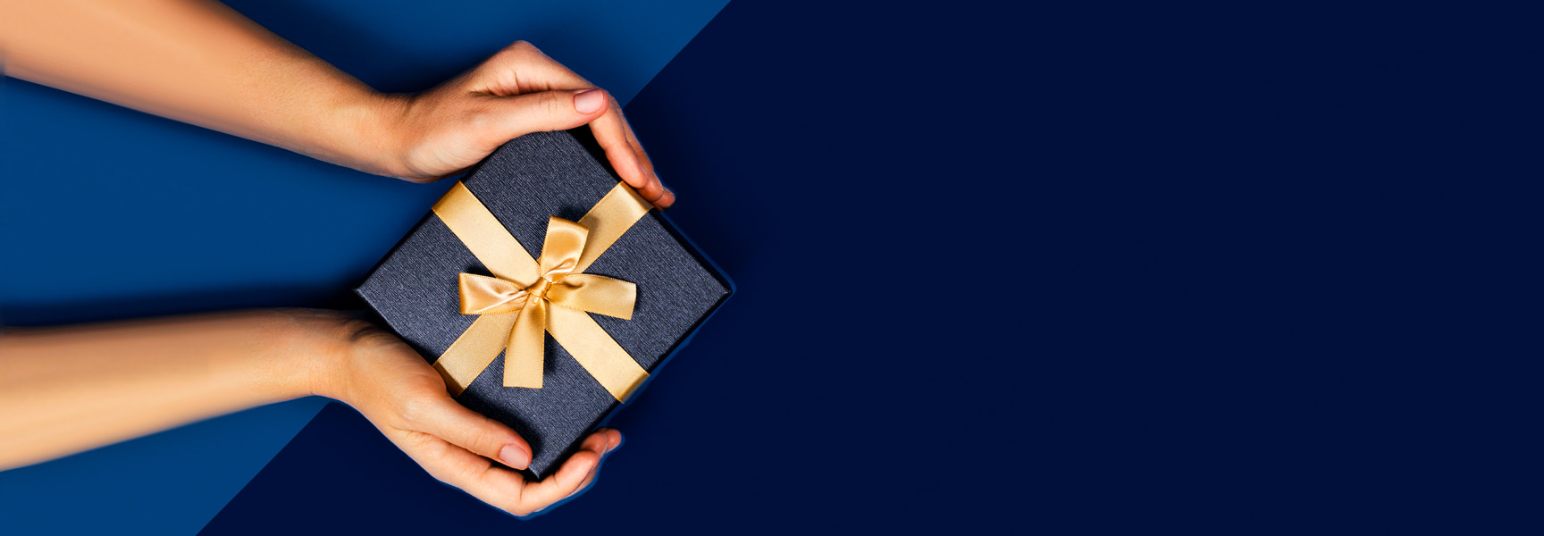 Hands holding a dark blue gift box tied with a gold ribbon bow, positioned on a blue background with empty space on the right.
