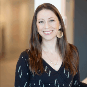 A woman with long brown hair smiles at the camera, wearing a black patterned top, large round gold earrings, and a delicate necklace. The background is softly blurred, suggesting an indoor setting.