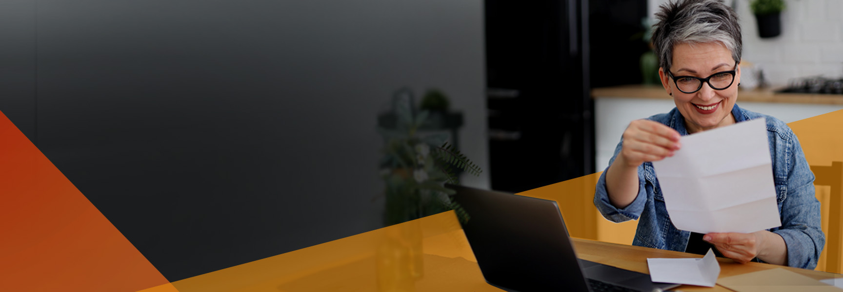 A smiling person with short gray hair and glasses sits at a table, holding up a piece of paper while looking at a laptop in a bright, modern home setting.