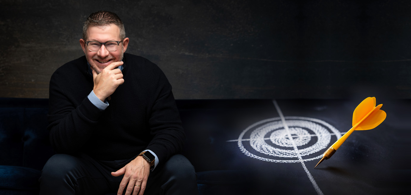 A man wearing glasses and a black sweater sits on a dark sofa, smiling with his hand on his chin. Next to him is a chalk-drawn target with a yellow dart hitting the bullseye.