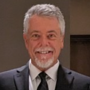 A man with short gray hair and a beard smiles while wearing a dark suit, white shirt, and black tie, standing indoors in front of a neutral background.