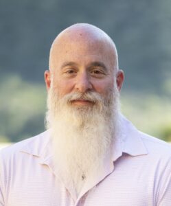 A man with a bald head and a long white beard wears a light-colored collared shirt and stands outdoors with greenery blurred in the background.