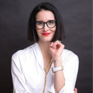 Woman with chin-length black hair and glasses, wearing a white shirt and red lipstick, poses confidently with her hand near her face against a dark background.