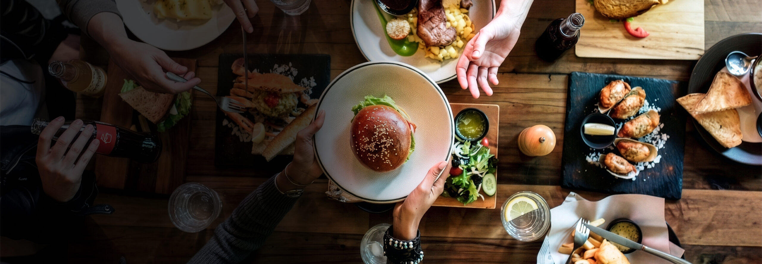 People sitting around a wooden table with various dishes, including a burger, wings, salad, steak, and drinks, reaching for food and sharing a meal together.